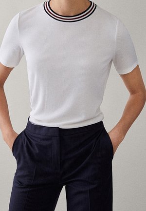 White ribbed t-shirt with a round neckline featuring a black and red striped trim; paired with navy striped pants, hands in pockets.