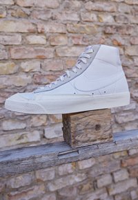 Valkoiset korkeajalkaiset nahkakengät, joissa on teksturoidut yksityiskohdat, tasaiset nauhat ja kumipohja, esiteltyinä puupalkilla tiiliseinän edessä.