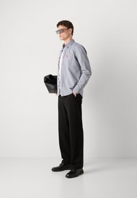 Cămașă bleu deschis cu butoni, cu un buzunar, asortată cu pantaloni negri și pantofi negri. Modelul ține o geantă neagră și poartă ochelari de soare.