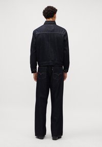 Temno modra denim jakna in usklajeni široki hlače z rumeno nitjo. Zadnji pogled prikazuje enostavno zasnovo in dva zadnja žepa z logotipom.