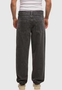 Man in losse donkergrijze jeans en beige sneakers, staand met zijn rug naar de camera op een effen lichte achtergrond.