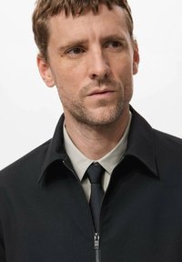 Črni blazer z zadrgo in ovratnikom, nošen preko svetle srajce in kravate. Značilnosti vključujejo gladko teksturo in srebrno zadrgo.