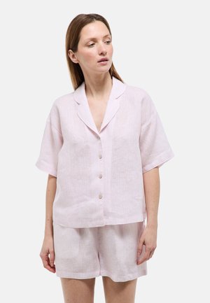 Femme portant une chemise à manches courtes rose clair à boutons et un short assorti, debout et regardant légèrement vers sa gauche.