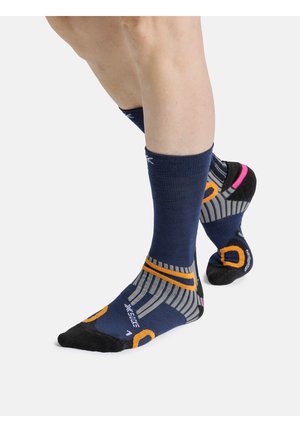 Persoon draagt marineblauwe, zwarte en oranje sportsokken en staat met één voet lichtjes geheven tegen een witte achtergrond.