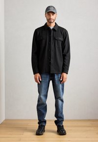 Mand iført sort skjortejakke, blå jeans, sorte sneakers og grå kasket står mod en ensfarvet væg på trægulv.