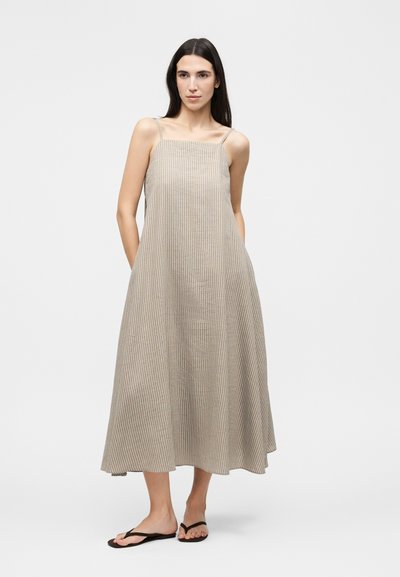 Γυναίκα με μακριά σκούρα μαλλιά φοράει ένα μπεζ και λευκό ριγέ αμάνικο μεσαίο φόρεμα και μαύρα σαγιονάρες, στέκεται με τα χέρια στις τσέπες.