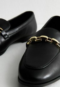Mocasines de cuero negro con un acento de cadena dorada en la parte frontal, con una textura suave y forma de puntera ovalada.