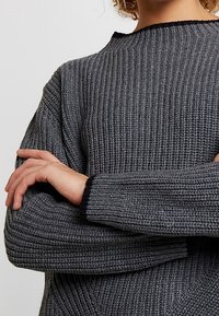 Pull en maille grise avec une texture côtelée et des accents noirs au niveau de l'encolure. Manches longues croisées devant, mettant en valeur l'épaisseur du tissu.