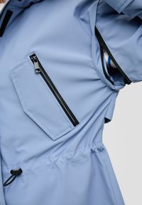 Jasnoniebieska kurtka wykonana z miękkiego materiału, z ukośną kieszenią zapinaną na zamek i regulowanym sznurkiem w talii. Czarny akcent zamka.