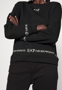 Zwarte sweatshirt met lange mouwen, voorzien van gouden EA7 Emporio Armani-logo's op de borst en zoom. Zacht materiaal; relaxed fit.