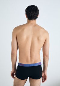Schwarze Boxershorts mit einem elastischen Bund in Blau und Rot, auf dem "HUGO" steht. Glattes Material und körpernahe Passform. Rückansicht des Modells.