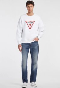 Balta džemperis su raudonos ir juodos spalvos trikampiu logotipu, kuriame yra užrašas „GUESS“ ir klaustukas, derinamas su mėlynais džinsais ir baltais sportbačiais.