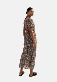 Maxi-jurk van doorzichtig bruin materiaal met een luipaardprintpatroon. Heeft korte mouwen, een uitsnijding op de rug en een zijsplit. Gekleed met zwarte sandalen.