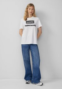 Weißes Baumwoll-T-Shirt mit schwarzem "Oasis Supersonic"-Aufdruck, kombiniert mit blauen ausgestellten Jeans. Sneakers sind unten sichtbar.