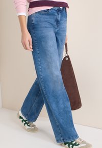 Femme portant un jean large bleu, un pull rose, des baskets blanches avec des détails verts, et tenant un sac en daim marron en marchant.