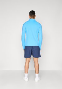 Ljusblå långärmad polo, marinblå shorts, vita sneakers och vita strumpor. Slät textur, avslappnad design och åtsittande silhuett, sedd bakifrån.