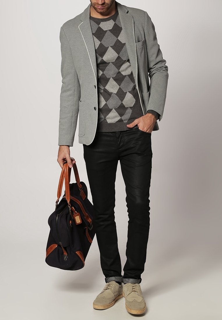 Homme en blazer gris et pull à motifs tenant un sac noir et marron avec des accents en cuir, portant un pantalon noir et des chaussures beiges.