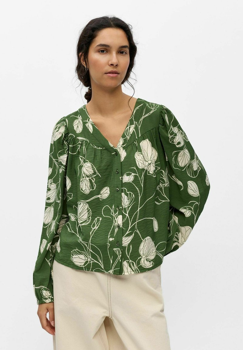 Femme portant un chemisier vert boutonné avec des motifs floraux blancs et un pantalon couleur crème, debout devant un fond uni.