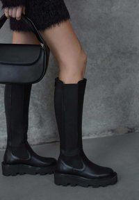 Black knee-high boots featuring a smooth leather toe, textured fabric shaft, thick sole with a treaded design, and a black handbag.