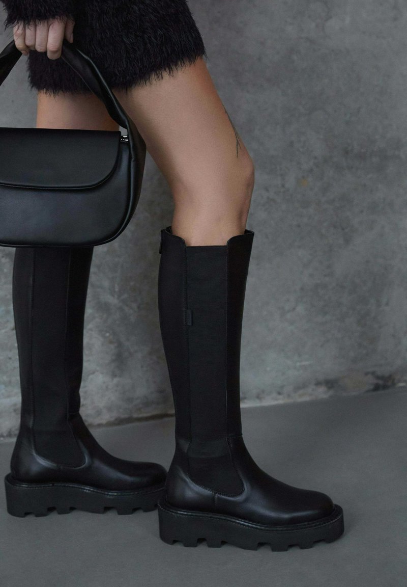 Black knee-high boots featuring a smooth leather toe, textured fabric shaft, thick sole with a treaded design, and a black handbag.