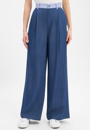Wide-leg blue denim trousers with pleats, a striped waistband, button closure, worn with white sneakers and a white top.