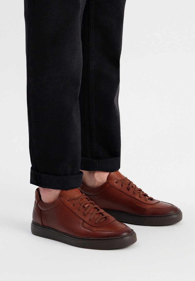 Braune Ledersneaker mit glatter Textur, runder Zehenpartie und Gummisohle. Kombiniert mit schwarzen umgekrempelten Jeans, die ein lässiges Design zeigen.