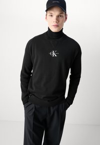 Black knit sweater with a high collar and textured sleeves. Features a white logo at the chest. Paired with dark trousers and a cap.