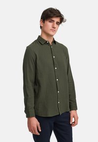 Olivgrüne Hemdjacke mit strukturierter Stoffqualität, langen Ärmeln und einem Kragen. Verfügt über weiße Knöpfe entlang der Vorderseite.