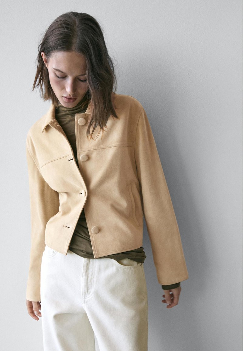 Beige ruskindjakke med et enkelt design, der har krave og knapper foran, kombineret med en brun højhalset trøje og hvide bukser.