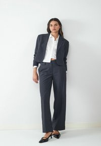 Marineblauw maatpak met een verkorte blazer en rechte broek. Witte blouse eronder, gecombineerd met zwarte hakken.