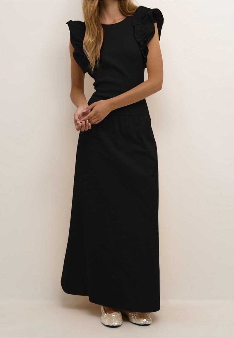Femme portant une longue robe noire à manches courtes volantées et des ballerines argentées, debout devant un fond clair uni.