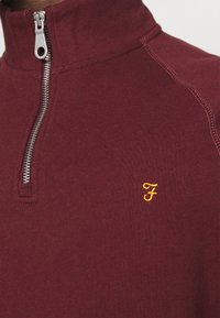 Burgundská mikina na zip s vysokým límcem, stříbrným zipem a malým žlutým vyšitým logem na hrudi; hladká textura látky.