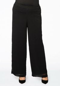 Zwarte wijde broek gemaakt van gestructureerde stof, met een comfortabele elastische tailleband en doorzichtige zoomaccenten. Gecombineerd met zwarte hoge hakken.