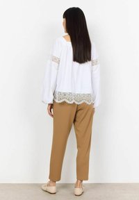 Hvid bluse med blonde detaljer og bølget nedre kant, parret med beige bukser. Modellen har langt mørkt hår og ses fra ryggen.