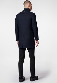 Ciemnoniebieski płaszcz wełniany z wysokim kołnierzem, pojedynczym tylnym rozcięciem i dopasowanym krojem. Noszony na wąskich czarnych spodniach i masywnych czarnych butach.