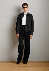 Person wearing sunglasses, black pinstripe jacket, white turtleneck, black wide-leg pants, and black shoes, standing against gray wall.