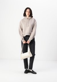 Sudadera gris recortada con logotipo, jeans negros y mocasines negros; sosteniendo un bolso rectangular beige claro. Diseño simple, estilo casual.