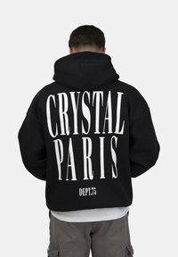 Sudadera negra con texto blanco en la parte posterior: "CRYSTAL PARIS" y "DEPT.75." Presenta una gran capucha y un corte relajado, confeccionada en tela suave.