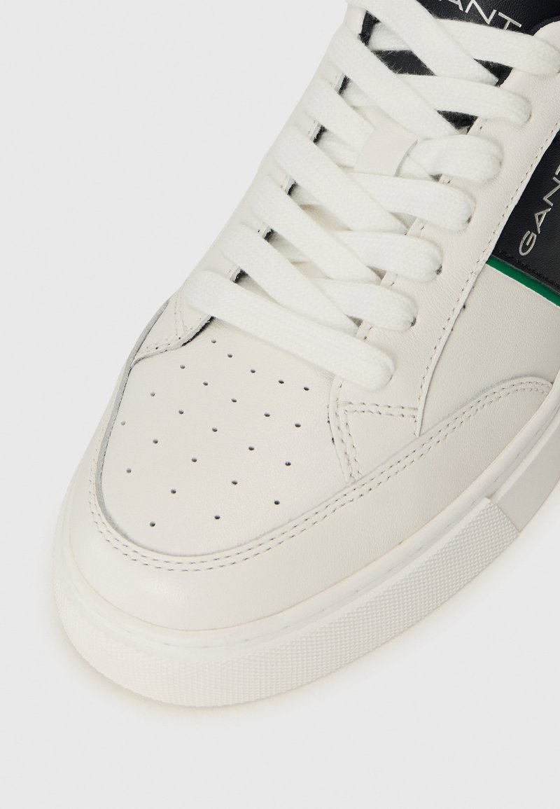 Weiße Sneaker aus glattem Leder mit durchbrochener Zehenbox, flachen weißen Schnürsenkeln und seitlichen Akzenten in Schwarz und Grün. Strukturiierte Sohle.
