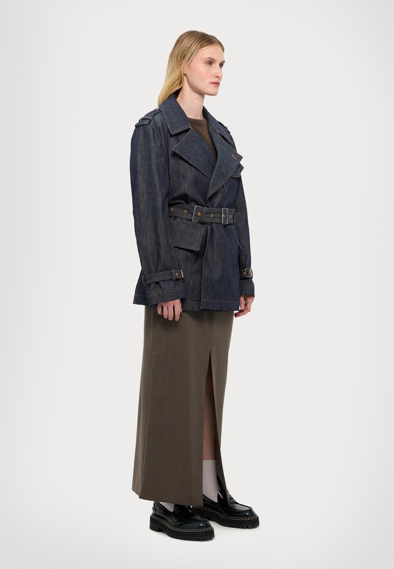 Manteau trench en denim avec taille ceinturée, design croisé, large col et épaulettes. Porté sur une longue jupe olive et des chaussures noires.