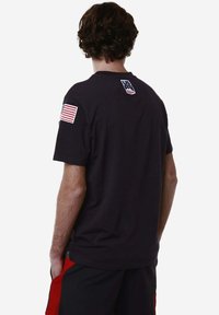 T-shirt nero a maniche corte con una patch della bandiera americana sulla manica sinistra e un logo nella zona del collo sul retro. Realizzato in tessuto morbido.