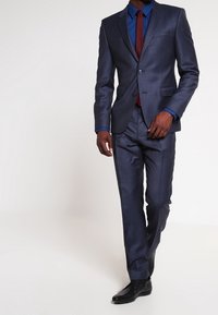 Marineblauer Anzug mit schlanker Passform, bestehend aus einer Jacke und passenden Hosen. Das Hemd ist blau, kombiniert mit einer burgunderroten Krawatte und schwarzen, polierten Schuhen.