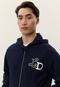 Homem jovem vestindo um hoodie azul-marinho com fecho de correr e logótipo "LD" no peito, posando contra um fundo claro e simples.