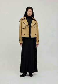 Lyhyt khaki-takki, jossa on kaulus ja etupainikkeet, pukeutuneena mustan poolopaidan ja pitkän mustan hameen päälle. Mustat nilkkakumit täydentävät asua.