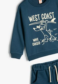 Marineblåt sweatshirt og matchende shorts med cremefarvede grafik af en hund med et surfbræt, blødt stof, ribbede manchetter og snor i taljen.