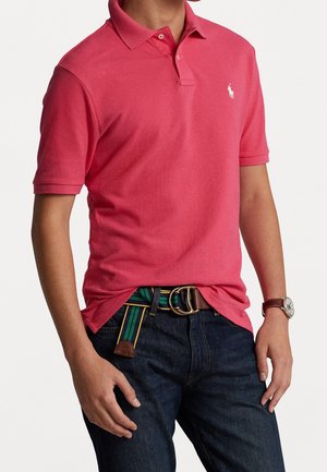 Homme portant un polo rose vif avec un jean sombre, une ceinture rayée verte et jaune, et une montre-bracelet au poignet gauche.