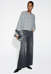 Grijze gebreide cardigan met knopen, gecombineerd met donkere wijde jeans. Model houdt een glanzende zilveren tas vast en draagt zwarte puntige schoenen.
