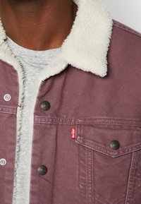 Burgundy denimjacka med en vit, fluffig krage. Funktioner inkluderar metallknappar, en bröstficka och en röd Levi's-logotypetikett.