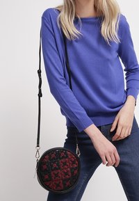 Czarna, okrągła torebka na ramię z czerwonym haftem kwiatowym, z teksturowaną powierzchnią i regulowanym paskiem. Torebka jest kompaktowa i stylowa.