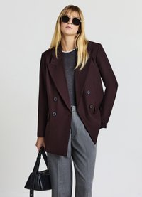 Burgundy double-breasted blazer with notch lapels, black buttons; paired with a gray top and tailored gray trousers; carrying a black handbag.